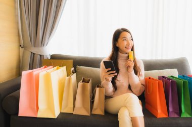 Portre güzel genç Asyalı kadın dizüstü bilgisayar ya da akıllı cep telefonu kullanarak oturma odasındaki alışveriş çantasının etrafındaki kanepede alışveriş yapıyor.