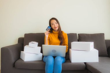 Asyalı genç bir kadının portresi, oturma odasındaki koltukta paket kutusuyla çalışmak için dizüstü bilgisayar kullanıyor.