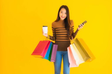 Güzel, genç Asyalı bir kadın sarı izole arka planda kahve fincanı ve renkli alışveriş çantasıyla akıllı cep telefonu veya cep telefonu kullanır.