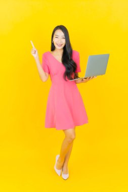 Asyalı güzel bir kadının portresi. Sarı izole edilmiş arka planda dizüstü bilgisayarla gülümsüyor.