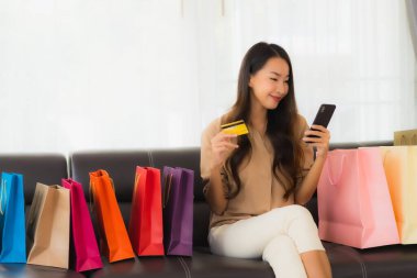 Güzel Asyalı bir kadının portresi. İnternetten alışveriş yapıyor. Kredi kartı ve akıllı telefonuyla oturma odasındaki alışveriş çantasının etrafında.