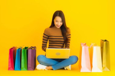 Güzel bir portre genç Asyalı kadın dizüstü bilgisayarla ya da dizüstü bilgisayarla yerde oturuyor sarı arka planda alışveriş torbası etrafında online alışveriş yapmak için kredi kartı kullanıyor.