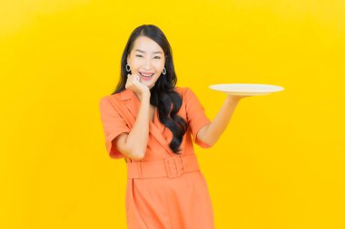 Asyalı güzel bir kadının portresi. Sarı arka planda boş tabak tabağıyla gülümsüyor.