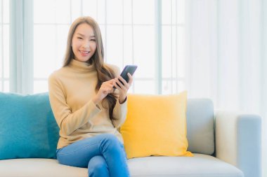 Oturma odasındaki kanepede akıllı cep telefonlu güzel Asyalı kadın portresi.