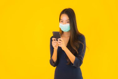 Maskeli ve akıllı cep telefonlu güzel bir Asyalı kadın portresi veya izole edilmiş sarı arka planda cep telefonu.