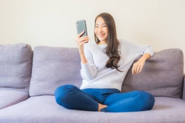 Güzel bir Asyalı kadın oturma odasındaki koltukta akıllı cep telefonu kullanıyor.