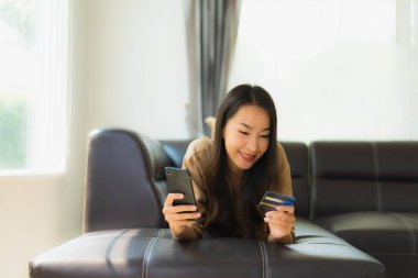 Akıllı telefon ya da kredi kartıyla oturma odasında online alışveriş yapan Asyalı güzel bir kadının portresi.