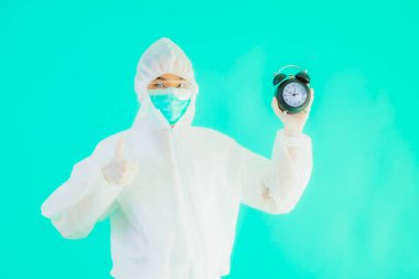Güzel bir portre genç Asyalı doktor. Özel süit ve maske takıyor. Mavi arka planda saati ya da alarmı gösteriyor.