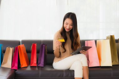 Güzel Asyalı bir kadının portresi. İnternetten alışveriş yapıyor. Kredi kartı ve akıllı telefonuyla oturma odasındaki alışveriş çantasının etrafında.