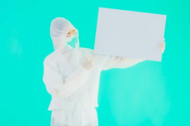 Güzel, genç Asyalı doktor kadın koronavirüsten korunmak için ppe ya da kişisel koruyucu ekipman takıyor ya da mavi izole arka planda beyaz kartı olan 19. bölgede yaşıyor.