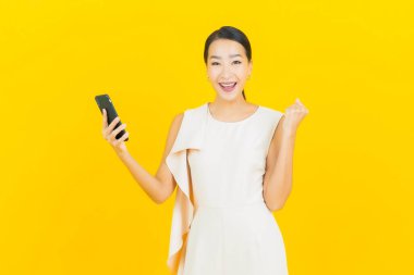 Asyalı güzel bir kadının portresi sarı arka planda akıllı cep telefonuyla gülümsüyor.