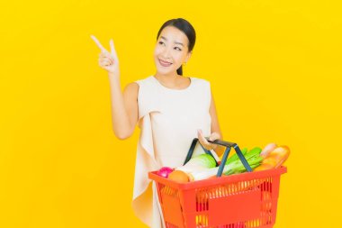 Asyalı güzel bir kadının portresi. Sarı arka planda süpermarketten alınmış alışveriş sepetiyle gülümsüyor.