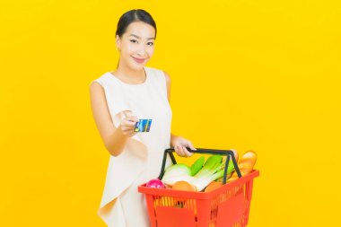 Asyalı güzel bir kadının portresi. Sarı arka planda süpermarketten alınmış alışveriş sepetiyle gülümsüyor.