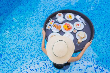 Asyalı güzel bir kadının portresi yüzme havuzunda yüzen kahvaltı tepsisiyle mutlu bir gülümseme