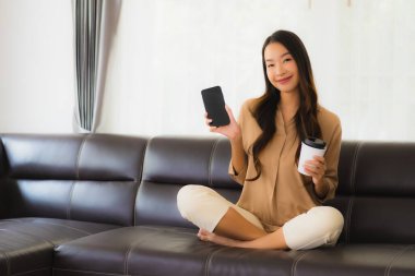 Güzel bir Asyalı kadın portresi. Kanepenin iç tarafında cep telefonu ya da kahve fincanı ile cep telefonu kullanıyor.