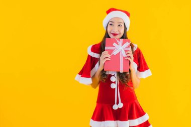 Güzel genç Asyalı noel kıyafetleri ve şapka gülümsemesi sarı izole edilmiş arka planda kırmızı hediye kutusuyla mutludur.