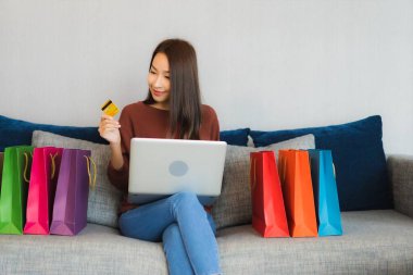 Asyalı güzel bir kadının portresi oturma odasındaki kanepede online alışveriş yapmak için bilgisayar ve kredi kartı kullanıyor.