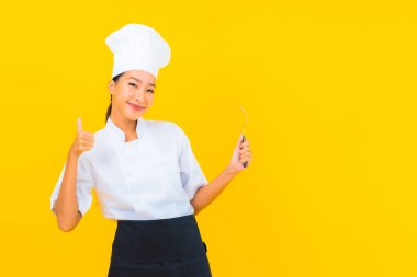 Güzel bir portre genç Asyalı kadın aşçı ya da aşçı üniforması giyiyor sarı izole arka planda çatal ve kaşıkla.