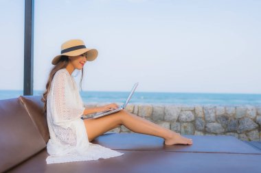 Plaj denizi çevresinde çalışmak için dizüstü bilgisayarı kullanan Asyalı genç bir kadının portresi.