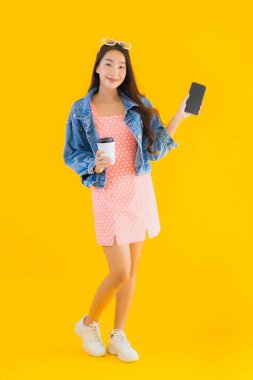 Kahve fincanı ve sarı izole edilmiş arka planda akıllı cep telefonlu güzel Asyalı kadın portresi.