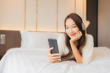 Güzel bir Asyalı kadın portresi. Yatak odasında müzik dinlemek için kulaklık ve akıllı cep telefonu kullanıyor.