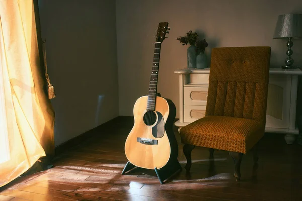 An acoustic Spanish guitar on a stand next to a chair in the moody shadows of a dark room with bright light coming in from behind a curtain