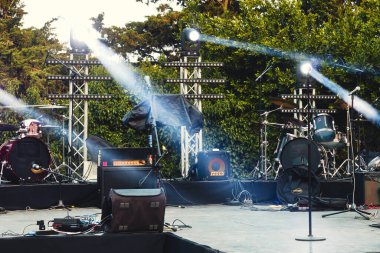 Gündüz vakti halka açık bir parkta hareketli spot ışıklarıyla aydınlatılmış, müzik ekipmanları ve gitar amfileri olan boş bir konser sahnesi.
