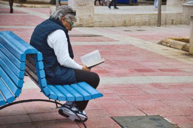 A middle-aged white Caucasian man sitting on a public park bench reading a book