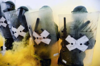 Riot police forming a line carrying perspex shields and wearing helmets - yellow smoke clipart