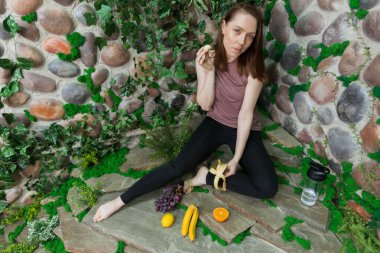 A healthy girl after yoga is eating fruits against the background of a overgrown stone wall. Concept of healthy food and lifestyle. Detox and clean diet.