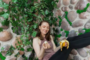 A healthy girl after yoga is eating fruits against the background of a overgrown stone wall. Concept of healthy food and lifestyle. Detox and clean diet.