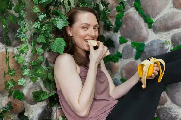 A healthy girl after yoga is eating fruits against the background of a overgrown stone wall. Concept of healthy food and lifestyle. Detox and clean diet.
