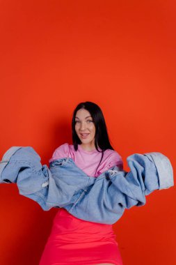 Close-up portrait of charming joyful beautiful girl posing with jeans against a bright red background. People emotions, lifestyle leisure, creative fashion concept.