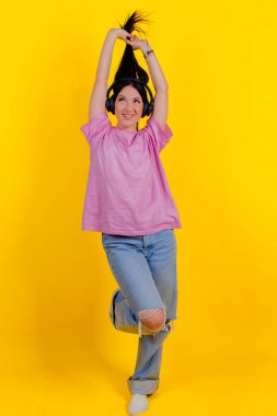 Portrait of a charming, joyful music lover holding, pulling pigtails or hair in her hands against a bright yellow background. Emotions, lifestyle, leisure, music, and dance concept.