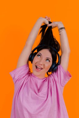 Portrait of a charming, joyful music lover holding, pulling pigtails or hair in her hands against a bright orange background. Emotions, lifestyle, leisure, music, and dance concept.