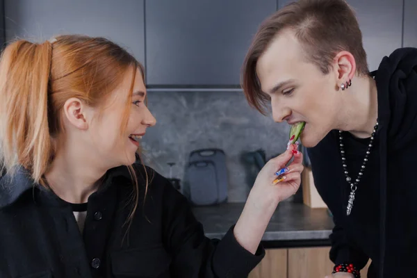 Young funny people spend time together in the kitchen. Girl feeds a guy a slice of cucumber.