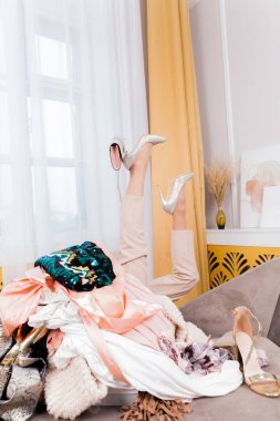 Woman legs out of a pile of clothes and accessories on the floor. Woman in high heels needs help from too much shopping. Shopaholic girl. Shopping addiction concept.