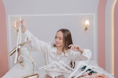 Young woman surrounded by different clothes and hangers in messy bedroom. many stacks of clothes. Disorder and mess. Clothing clutter and disorganization in the bedroom.