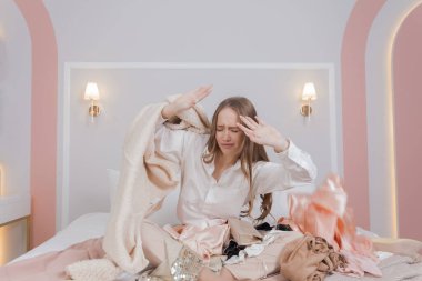 Attractive girl is sitting on a bed throwing clothes, a mess around. Nothing To Wear concept. Fashion style. Shopaholic is upset.