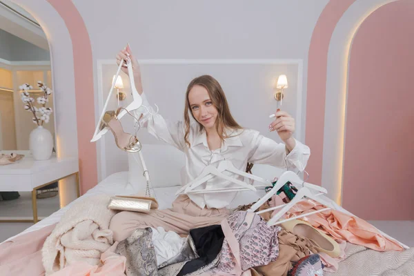 Young woman surrounded by different clothes and hangers in messy bedroom. many stacks of clothes. Disorder and mess. Clothing clutter and disorganization in the bedroom.