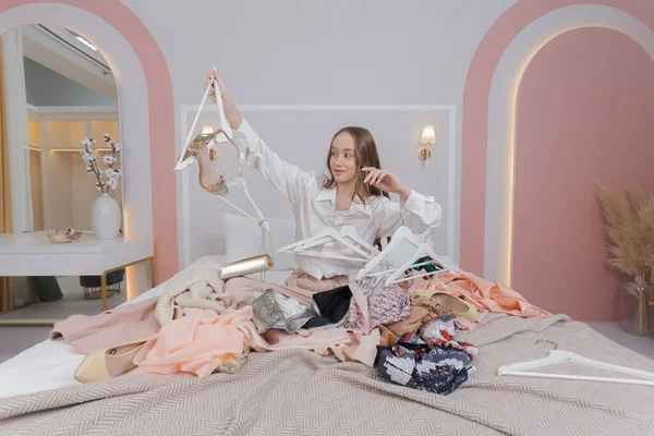 Young woman surrounded by different clothes and hangers in messy bedroom. many stacks of clothes. Disorder and mess. Clothing clutter and disorganization in the bedroom.
