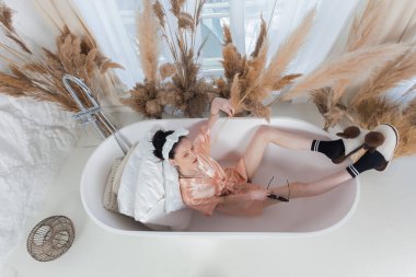 Attractive slim girl in a bathrobe with a bow on her head, wearing socks and slippers, is lying in a bathtub.