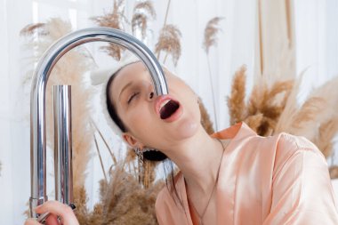 Attractive woman in bathrobe with a towel wrapped around her head opened her mouth drinking tap water over sink in kitchen or bathroom.