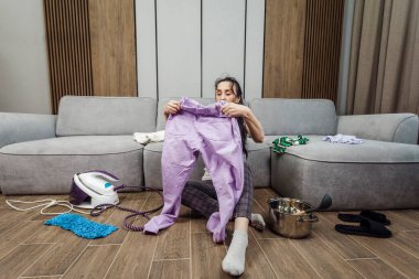 Tired middle age woman sitting on the floor with a pile of laundry and an iron.