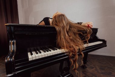Elegant young long haired brunette woman in black dress lying on a piano with hair falling down.