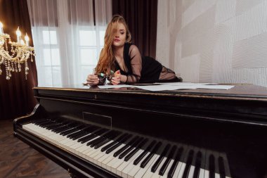 Elegant young long haired brunette woman in black dress lying on a piano holding glasses and looking at music sheets.