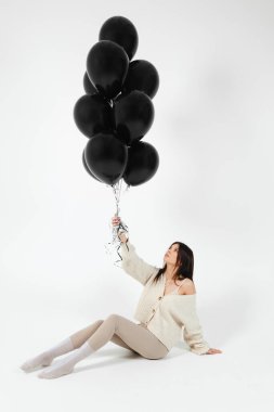 Brunette Girl in light clothes sitting on a floor and a Bunch of black balloons pushes her up.