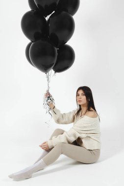 Brunette Girl in light clothes sitting on a floor and a Bunch of black balloons pushes her up.