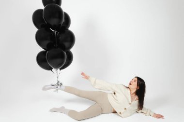 Brunette Girl in light clothes lying on a floor and a Bunch of black balloons pushes her up by leg.