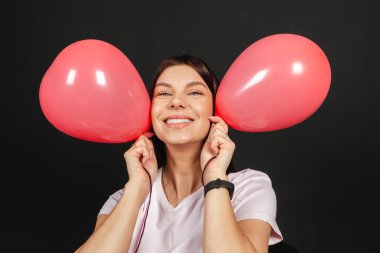 Portrait of emotional Young brunette in light clothing holding red balloons, isolated on black background.
